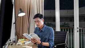 A man reading a book at a desk with a lamp, books, and a cup. - TreePark Karawang (East Telukjambe)