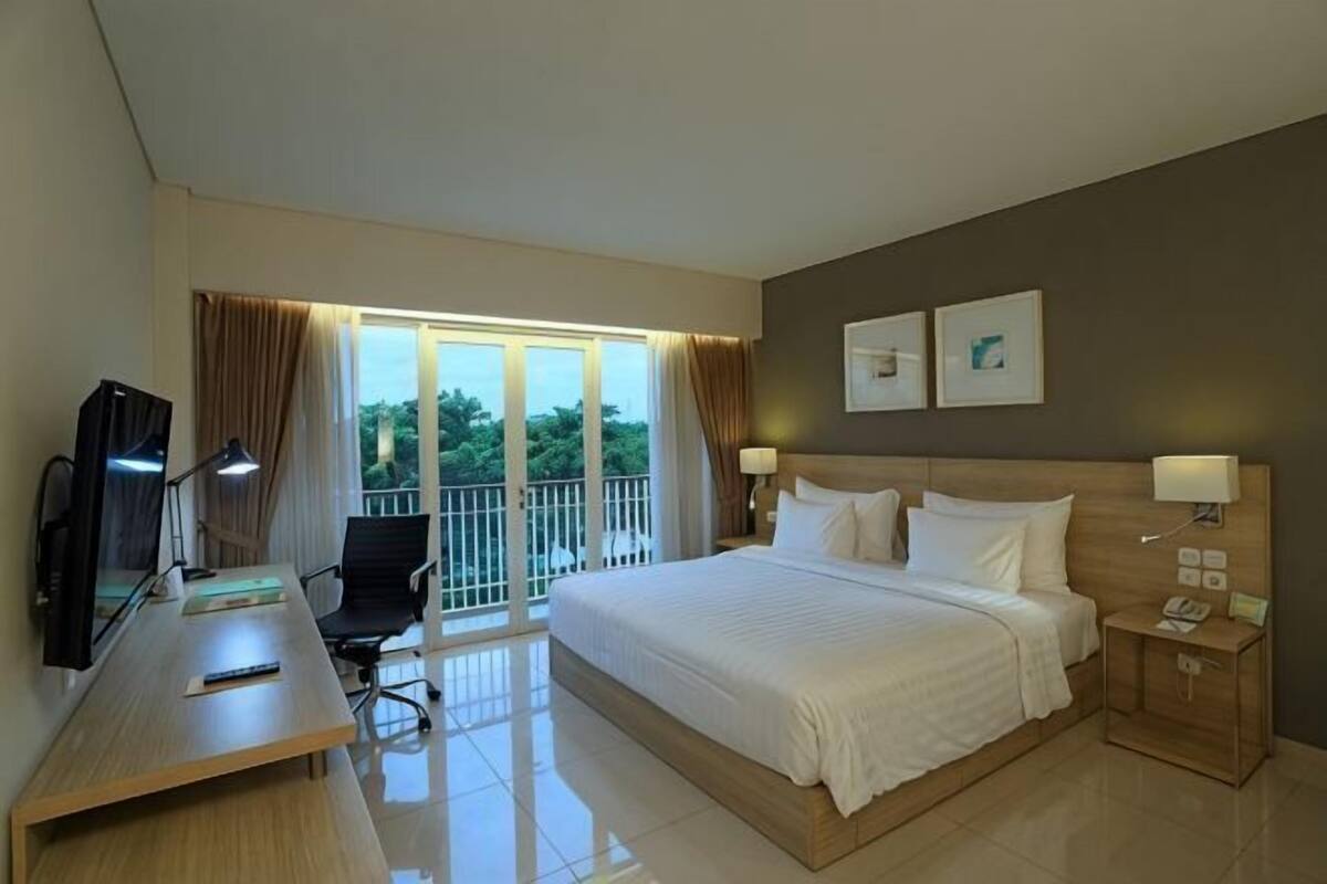 Habitación de hotel con cama, escritorio, silla, televisión y balcón con cortinas.