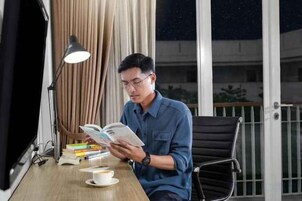 Un hombre leyendo un libro en una mesa con una lámpara, libros y una taza.