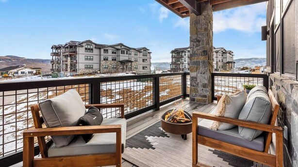 A balcony with two wooden chairs and a fire pit, overlooking a residential area with multiple buildings.