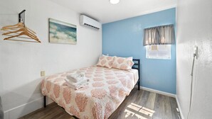 Un dormitorio con cama, perchero, aire acondicionado, ventana con cortinas e imagen colgada en la pared.
