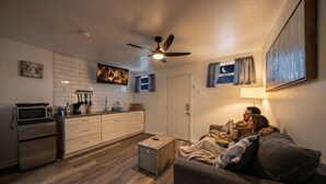 Una sala de estar acogedora con un sofá, un televisor, un ventilador de techo y una zona de cocina con armarios y electrodomésticos.