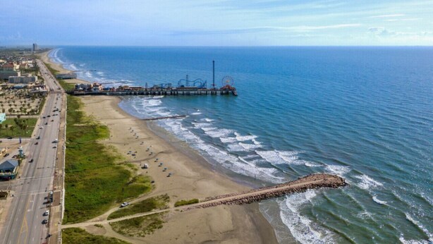 Vista aérea de una zona costera con playa, muelle y una carretera que recorre la orilla.