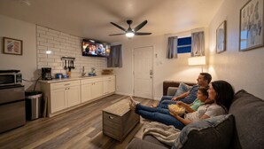Una sala de estar con un sofá, una televisión, un ventilador de techo y una cocina al fondo.