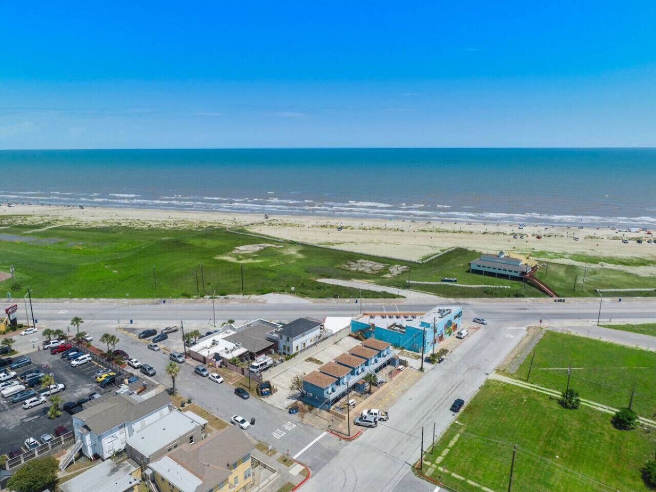 Vue aérienne d’une ville côtière avec une plage, un parking et des bâtiments, dont un immeuble bleu orné d’une fresque.