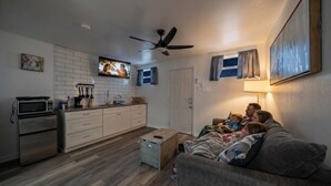Un salon confortable avec un canapé, un téléviseur, un ventilateur de plafond et un coin cuisine équipé de placards et d’appareils électroménagers.