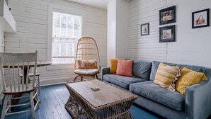 A room with a grey sofa, a wicker chair, a wooden coffee table, and a window with blinds. - Charming Historic Home by Beach & Strand (Galveston)