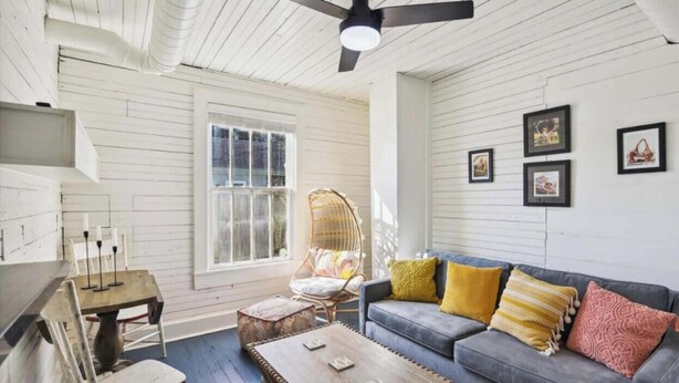 A cozy room with a gray sofa, a wicker chair, a wooden table, and a window with white shutters. - Charming Historic Home by Beach & Strand (Galveston)