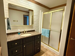 A bathroom with a white brick wall, a large mirror, a sink with a gold faucet, dark cabinets, and a shower with a glass door.