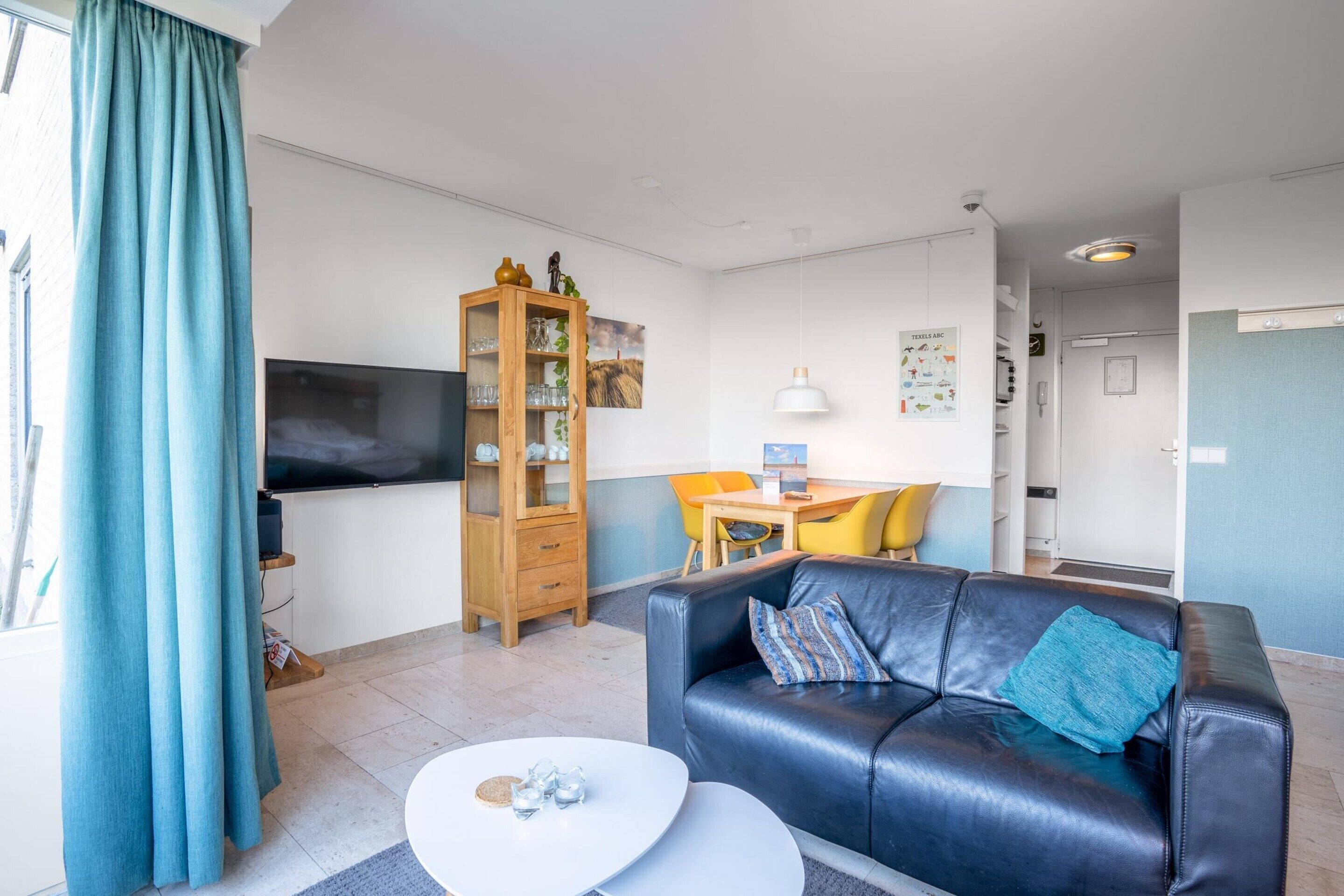 Una sala de estar moderna con un sofá de cuero negro, una mesa de centro blanca, un mueble de madera y un televisor de pantalla plana montado en la pared.