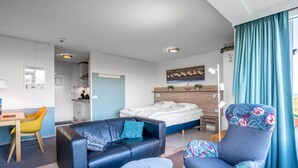 Una habitación moderna y luminosa con una cama, un sofá, una silla y una pequeña mesa. Se observa una zona de cocina al fondo.