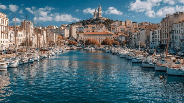 Un port esportiu amb vaixells amarrats al passeig marítim, edificis a ambdues bandes, i un turó destacat amb una torre al fons.