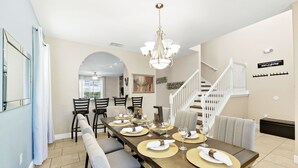 A dining area with a long table set for six, a chandelier, and a staircase leading to an upper level.