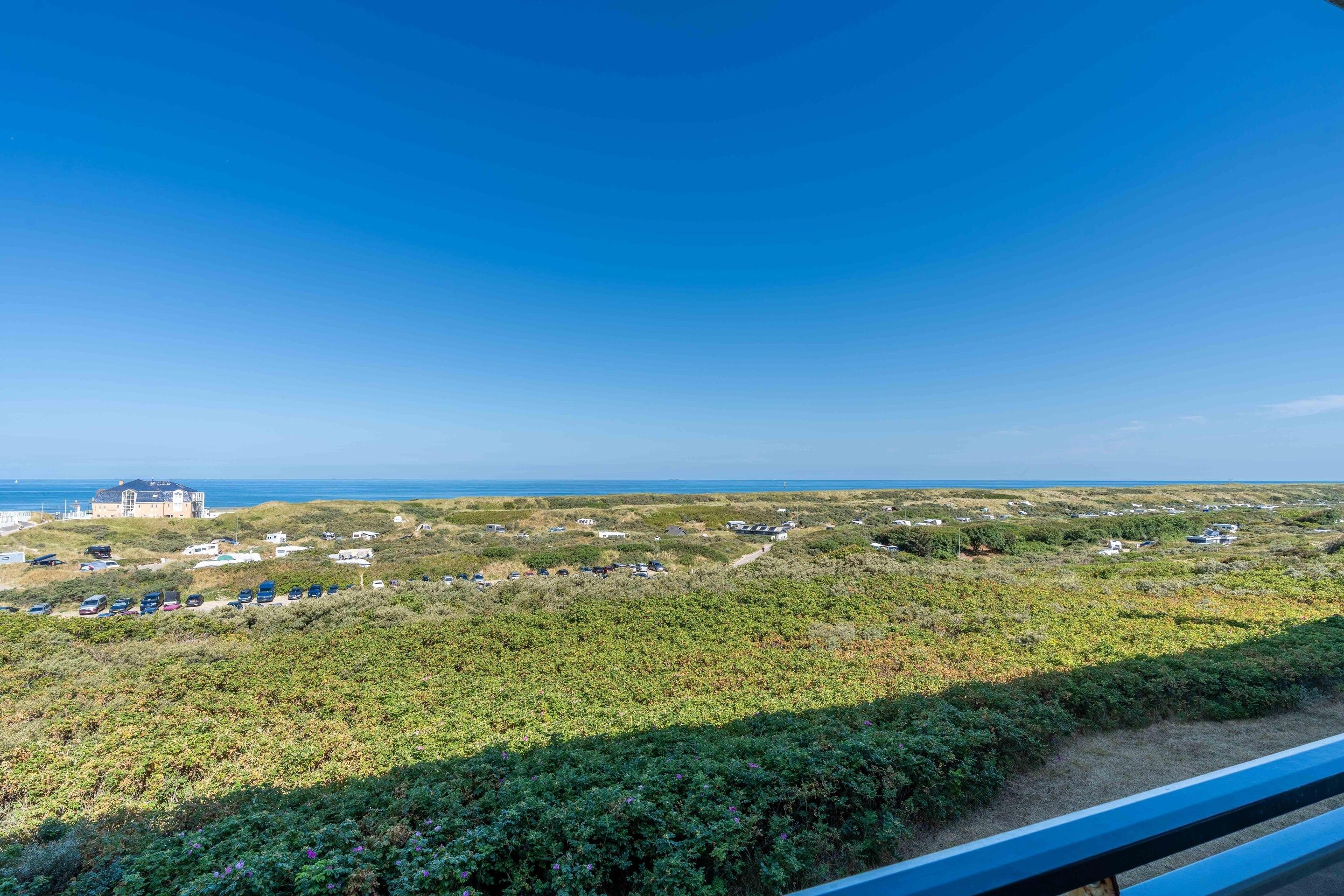 A coastal landscape with a view of the sea, a path leading through a grassy area, and several buildings in the distance.
