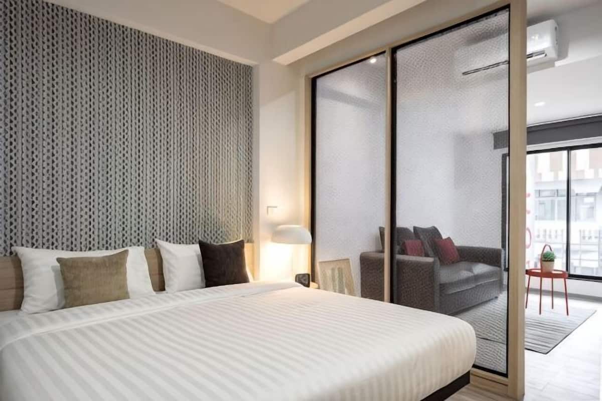 A modern hotel room with a large bed, a sofa, a small table, and a sliding glass door leading to a balcony.