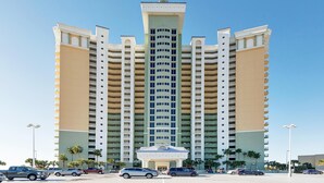Un hotel a più piani con una torre centrale, numerosi balconi e un parcheggio pieno di automobili.