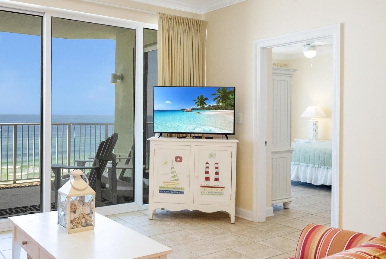 A room with a television on a cabinet, a view of the beach, a chair, and a bed with a white bedspread.
