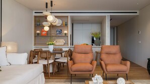 Una sala de estar moderna con un sofá, dos sillones, una mesa de comedor con sillas, una estantería y una zona de cocina al fondo.