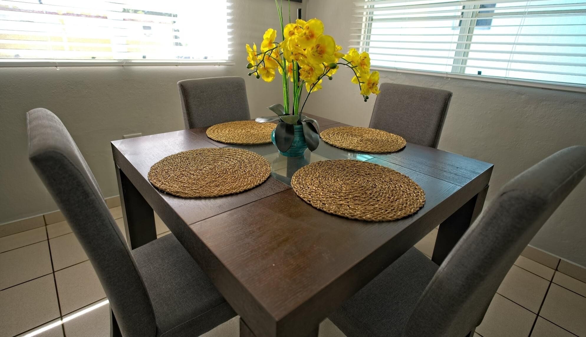 A dining table with four woven placemats, a vase with yellow flowers, and grey upholstered chairs.