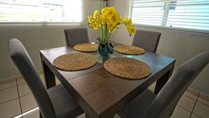 A dining table with four woven placemats, a vase with yellow flowers, and grey upholstered chairs. - Oceanfront Paradise: Private Balcony, Stunning Views & Steps to the Beach! (Aguadilla)