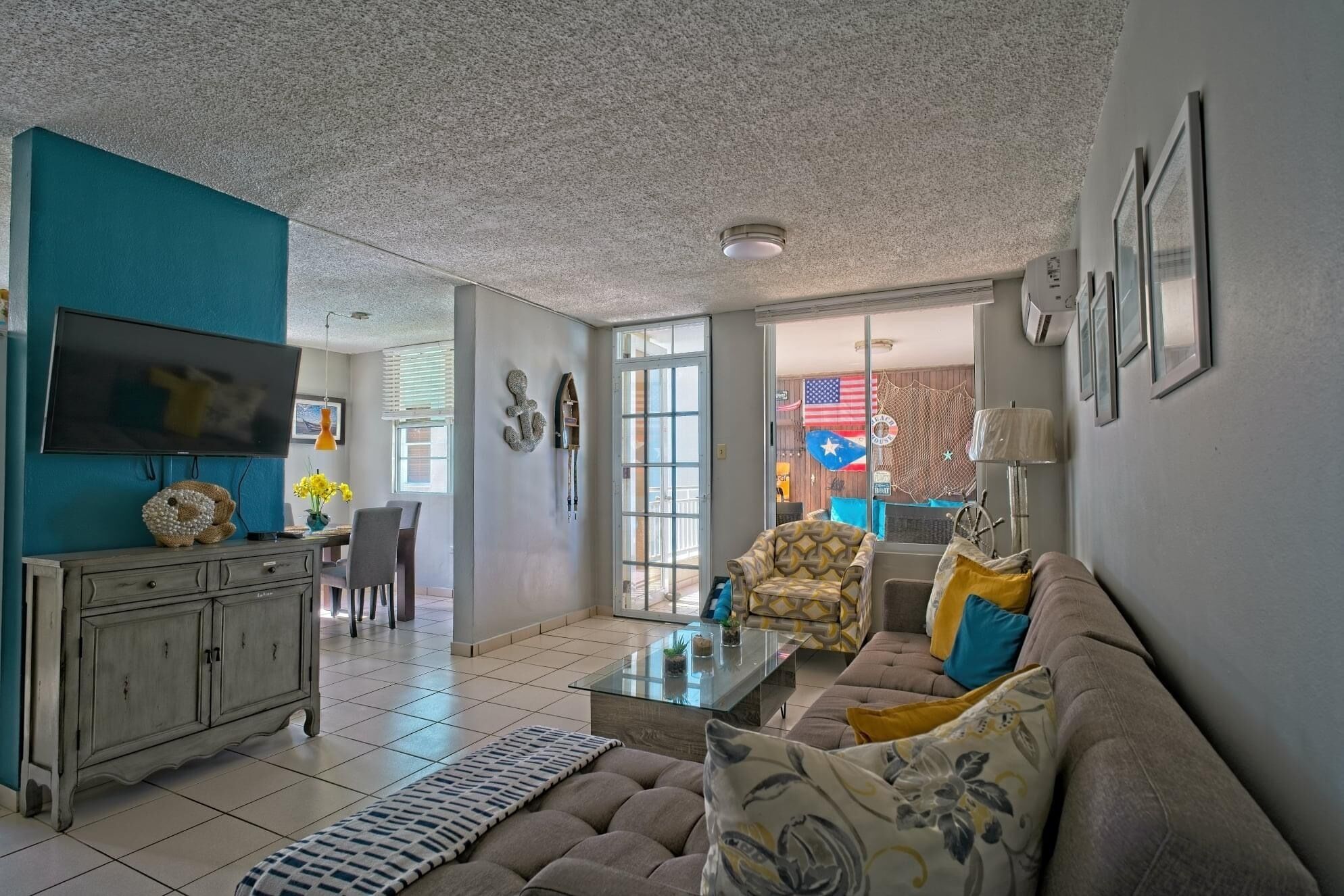 A living room with a sofa, coffee table, and a TV. There is a dining area with a table and chairs. The room has a ceiling with a textured finish and a door leading to another room.