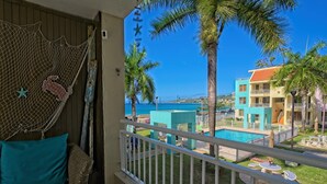 A balcony with a view of a pool, palm trees, and a building with a red roof. - Oceanfront Paradise: Private Balcony, Stunning Views & Steps to the Beach! (Aguadilla)