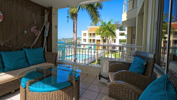 A balcony with wicker furniture, a glass table, and a view of a swimming pool and buildings. - Oceanfront Paradise: Private Balcony, Stunning Views & Steps to the Beach! (Aguadilla)