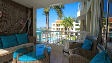 A balcony with wicker furniture, a glass table, and a view of a swimming pool and buildings.