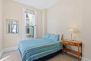 A bedroom with a bed, a bedside table with a lamp, a mirror, and a window with curtains.