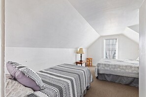 A small, white, attic-style bedroom with two beds, a window with blinds, a lamp, and a small table.