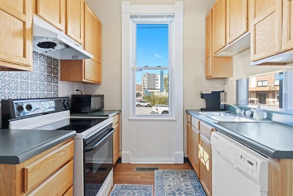 Cocina compacta con muebles de madera, microondas, estufa y lavavajillas. Una ventana ofrece vistas al exterior.