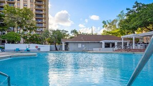 Una piscina con superficie de agua azul cristalina, rodeada por un edificio blanco con tejado de azulejos y un edificio residencial alto al fondo.