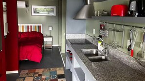 Een compacte keuken met een spoelbak, aanrecht en diverse keukengerei. Een bed met rode beddengoed is zichtbaar in de aangrenzende kamer.