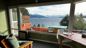 A room with a view of a lake and mountains, a wooden chair, a table with a glass, and a cushioned bench. - Loft with incredible lake views (San Carlos de Bariloche)