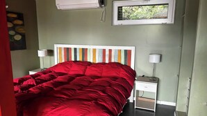Een slaapkamer met een rode sprei, een gestreept hoofdeinde en een raam met uitzicht op groen.
