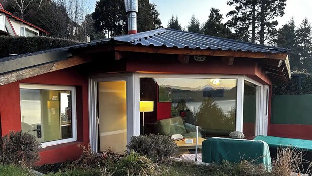 A small, single-story building with a red exterior and a metal roof. It features a large sliding glass door leading to an interior with a green table and a lamp. - Loft with incredible lake views (San Carlos de Bariloche)