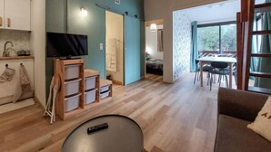 A modern living room with a TV, wooden storage units, a dining area with a table and chairs, and a large window offering a view of the outdoors.