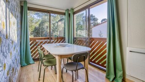 A dining area with a table and chairs, large windows with curtains, and a view of trees and a fence.