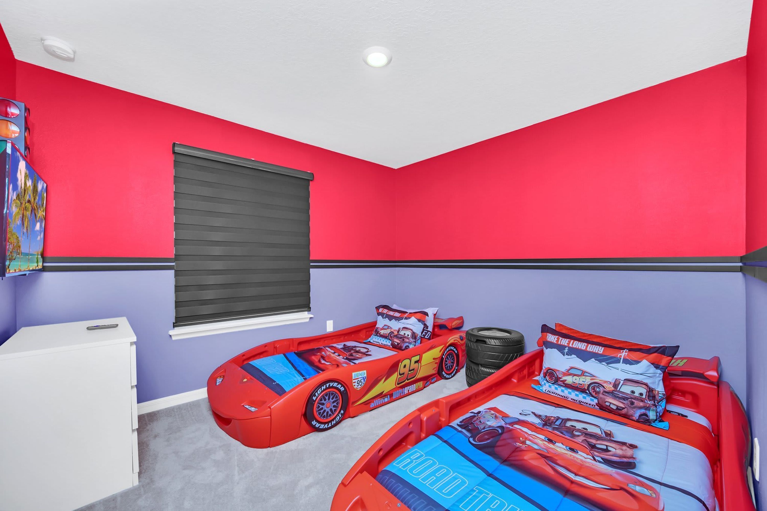 A bedroom with two beds, each with a 'Cars' themed bedspread, a white dresser, and a window with blinds.