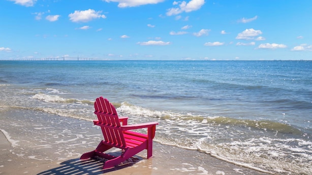 Une scène de plage avec une chaise Adirondack rose sur le sable, un ciel bleu clair et un pont au loin.