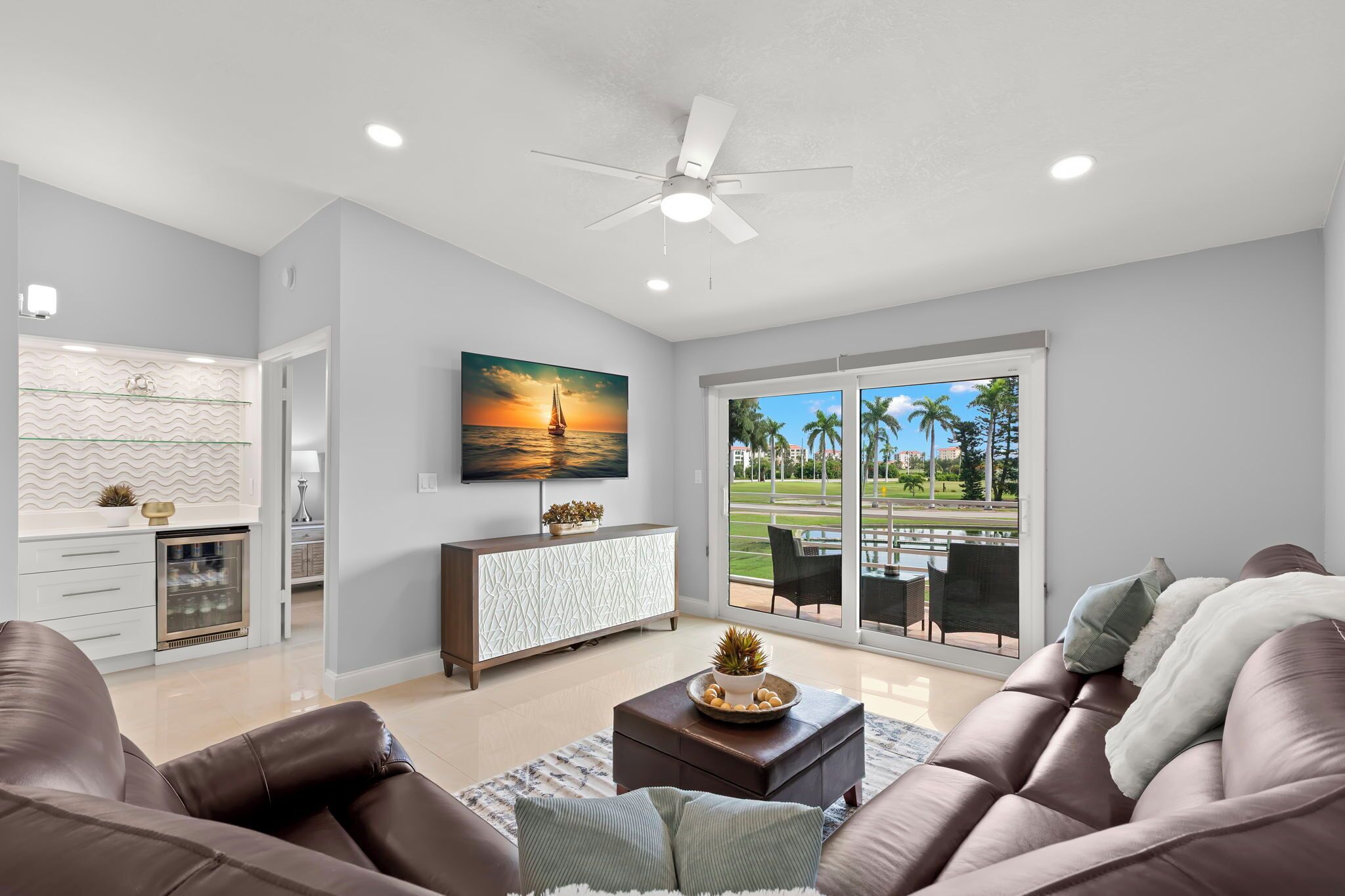 A modern living room with a brown leather sofa, a coffee table, and a television mounted on the wall. There is a large window with a view of palm trees and a patio.