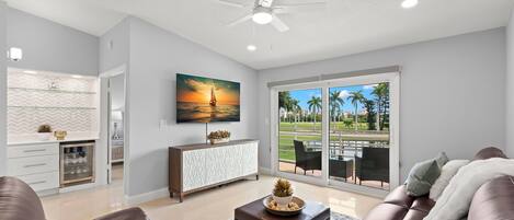 A modern living room with a brown leather sofa, a coffee table, and a television mounted on the wall. There is a large window with a view of palm trees and a patio.