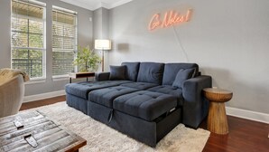 Un salón moderno con un sofá seccional gris oscuro, una alfombra blanca y esponjosa, una mesa de centro de madera, una mesita auxiliar y un letrero de neón en la pared.