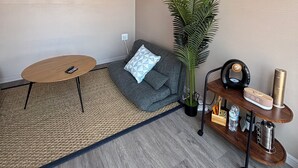 A modern living room with a grey sofa, a small round table, a large potted plant, a wooden side table with a clock and a water bottle, and a black projector mounted on the ceiling.