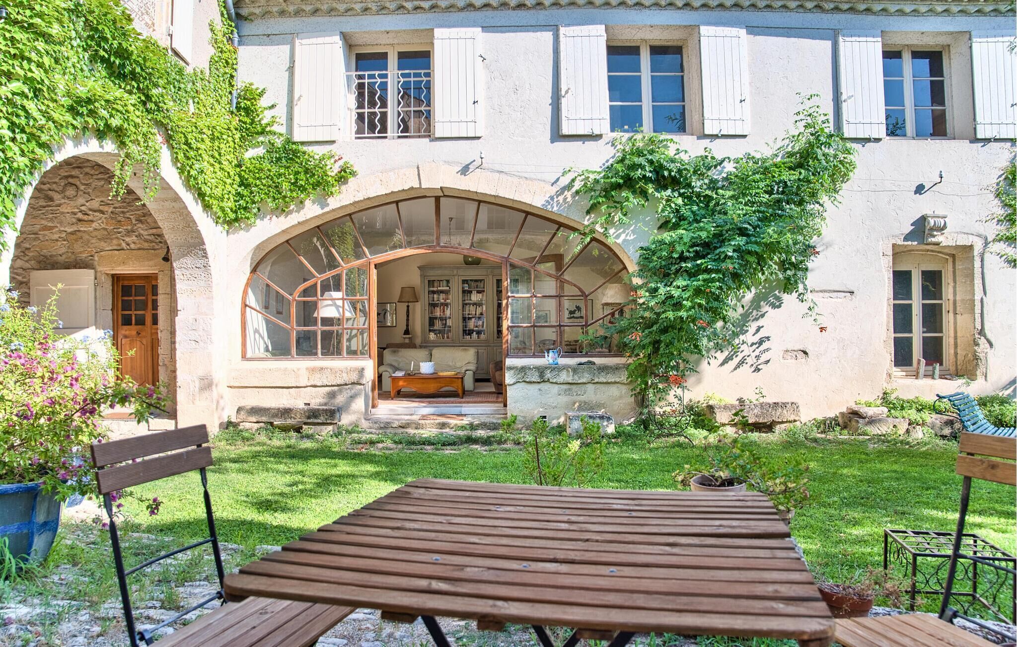 En bygning i sten med en overdækket terrasse, en træbænk og en have med plantekasser.