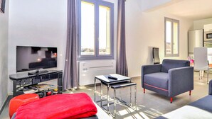 A modern living room with a flat-screen TV, a sofa, a small table, and a red ottoman. There is a window with curtains, a radiator, and a view of the outside. - Beautiful apartment in Uzès with WiFi (Uzès)
