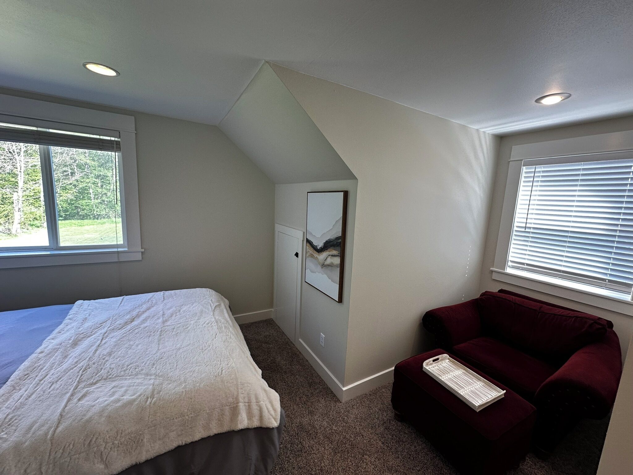 Une chambre à coucher avec un lit, une chaise rouge, un tableau encadré et une fenêtre avec des stores.