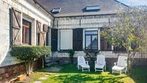 Ein Haus mit einem geneigten Dach, einem Ziegelkamin und einem kleinen Garten mit drei weißen Liegestühlen.