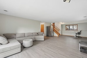 A spacious living room with a sectional sofa, a small ottoman, a refrigerator, a wooden staircase, and a window with blinds.
