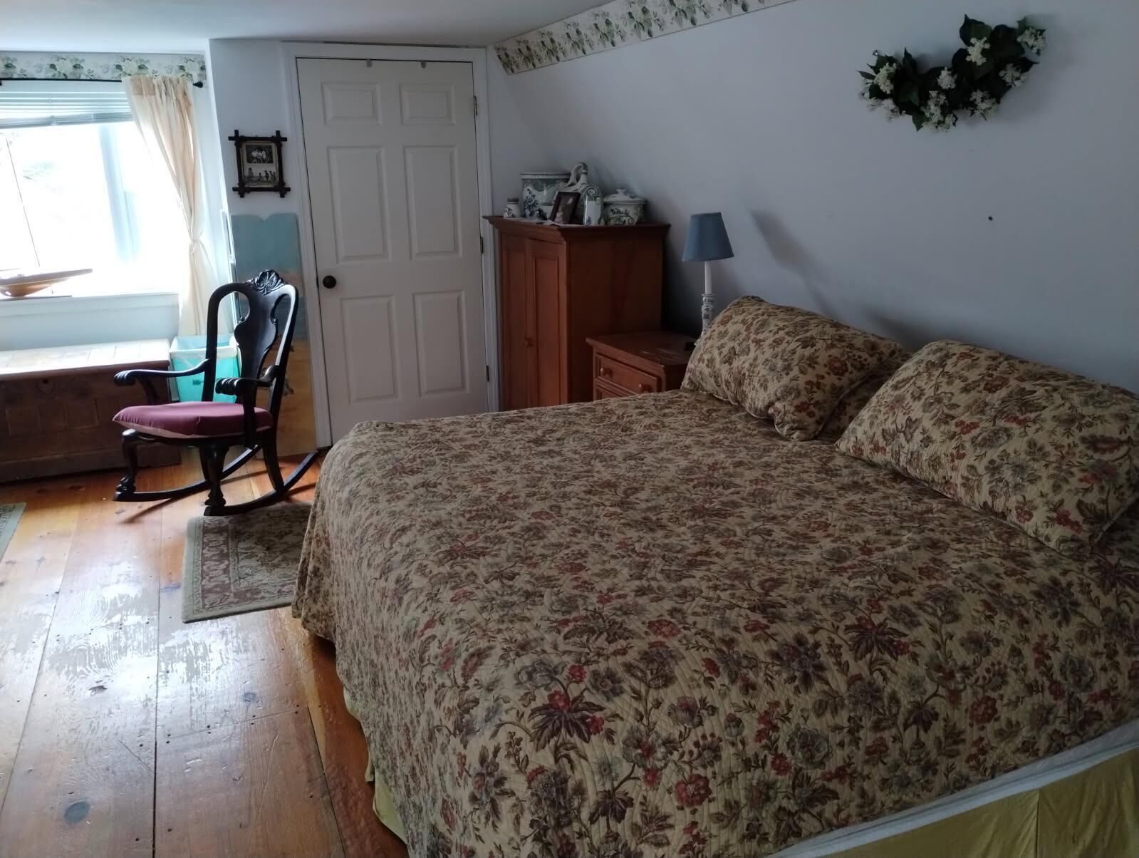 Ein Schlafzimmer mit geblümtem Bettbezug, einem Schaukelstuhl, einer Holzkommode und einer Lampe.